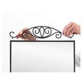 Two hands attach a decorative metal arch to a rectangular frame, likely the top of a mirror, enhancing its style and utility for hanging items.