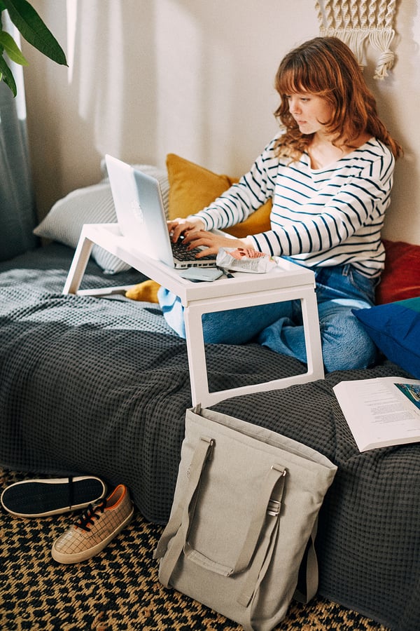 A woman works on a laptop on a KLIPSK white bed tray in bed with a striped shirt.
