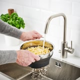 Person rinses pasta in stainless steel sink using black collapsible colander under modern silver faucet.