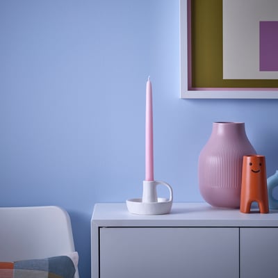 KLOKHET pink candle in white holder: small, bright, sits on cabinet next to vase, orange smiley figure, blue wall.