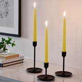Three bright yellow KLOKHET candles on black holders, lit, on white surface.