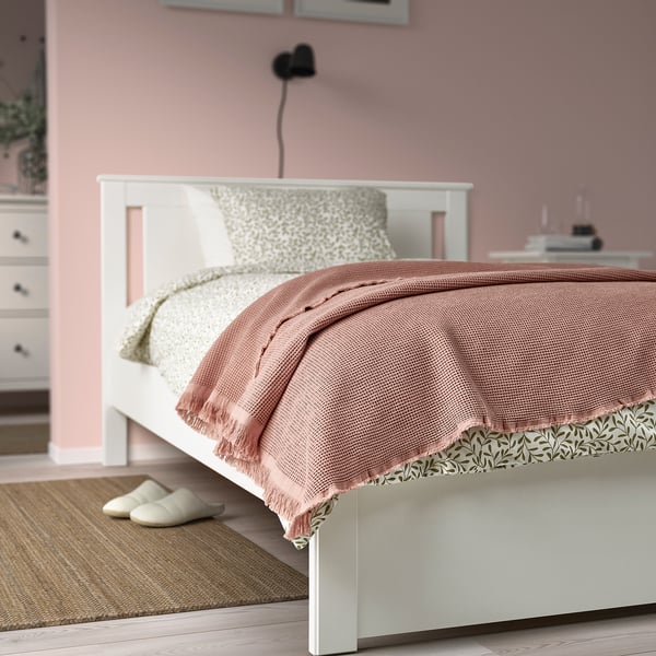 Cozy bedroom with pinkish-beige KLOTSTARR blanket, waffled texture, matching cushion, slippers, rug, and plant.