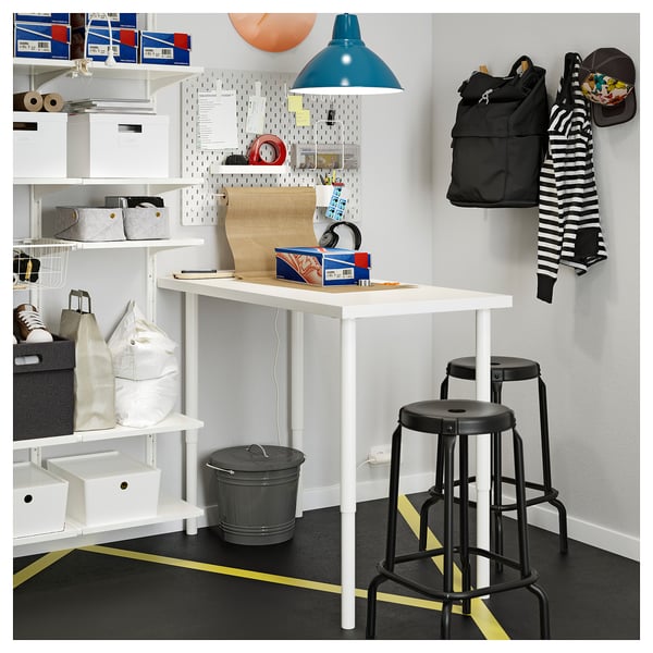 Well-organized workspace with white desk, two black stools, gray bin, shelves, and pegboard with various items.