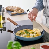 Person flips food in a blue pan using a KNORRHANE spatula. The spatulas wide blade and sloped handle aid in flipping and serving.