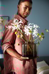 A person holds a transparent brown glass vase with white flowers and small yellow spheres, part of the KONSTFULL series designed by ilse crawford.