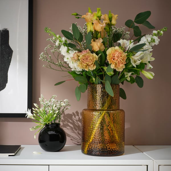 A KONSTFULL vase, brown, glass, cylindrical, designed to display flowers, larger than a small black vase beside it.
