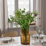A dining table with flowers in a brown textured glass vase, flanked by water glasses and plates on beige napkins, with wooden chairs.