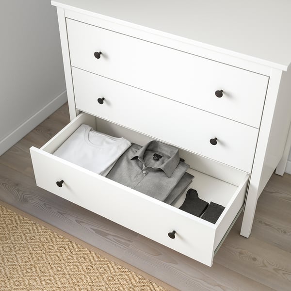 White chest of drawers with three open drawers revealing folded clothes. Safety wall anchoring indicated.