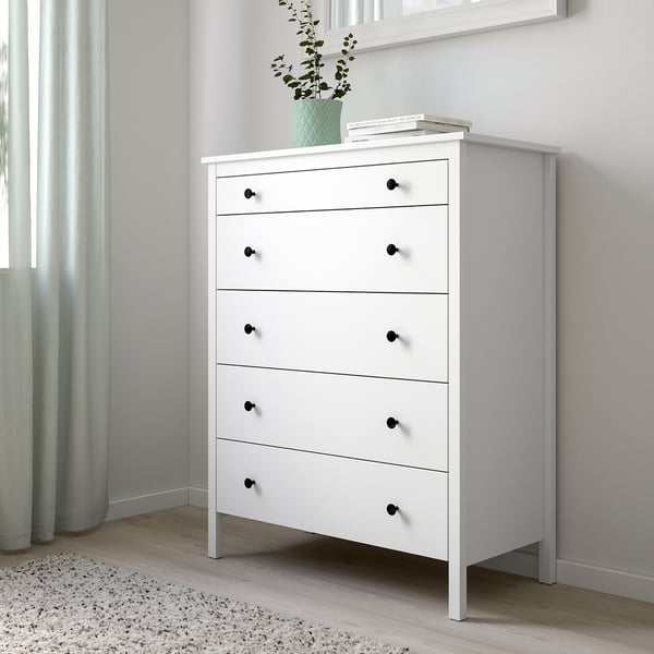 KOPPANG white chest with 5 drawers, black knobs, green plant, books, on rug, beside wall.