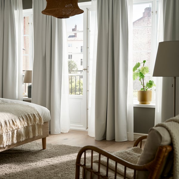 Bright bedroom with KORGMOTT white block-out curtains, a bed, chair, and plant. Curtains span floor-to-ceiling, framing a balcony door.