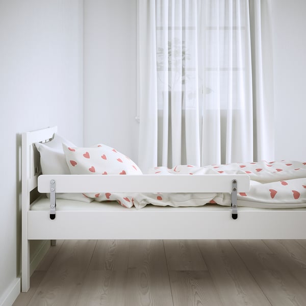 Small white kids bed with heart print sheets and guard rails by window.