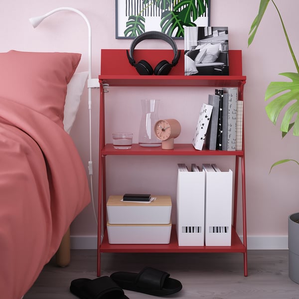 Red KRÖNIKÖR shelf beside bed, holding books, clock, headphones, lamp.
