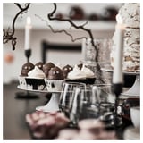 Ghostly cupcakes in KUSTFYR black & white cups sit on a cake stand, smaller than wine glasses nearby. Branches and candles decorate the table.