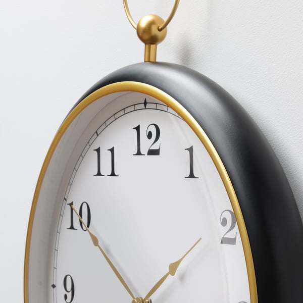A large, stylish wall clock with a matte black face and gold accents, featuring clear, large white numbers and gold hands.