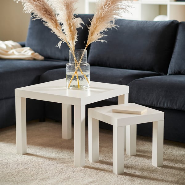 Two white LACK tables near a bLACK couch. The larger table holds a vase with flowers, while the smaller one holds a book.