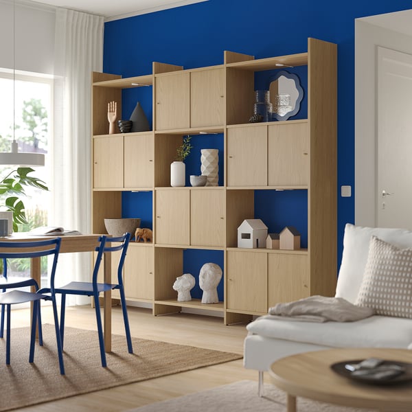 Modern living room: light wood bookshelf, blue accents, white couch, plants, natural light.