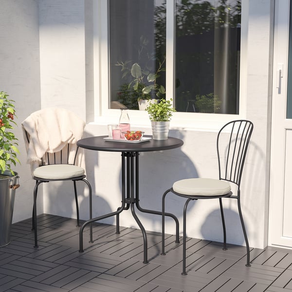 Outdoor table & chairs with cushions. Bowl of fruit, glasses, blanket. Patio behind glass door with greenery. Compact for small spaces.