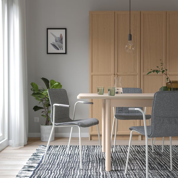 Modern dining area with wooden table, gray chairs, pendant light, plant, wood cabinets, rug, large window.
