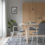 Modern dining area with wooden table, gray chairs, pendant light, plant, wood cabinets, rug, large window.