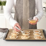 Person uses a brush from LÄTTBAKAD to glaze pastries on a tray.