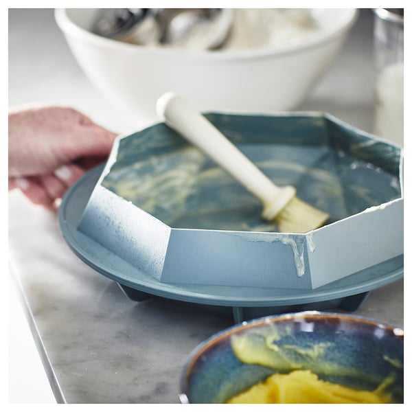 A blue LÄTTBAKAD baking brush being used to coat a cake in a blue pan.