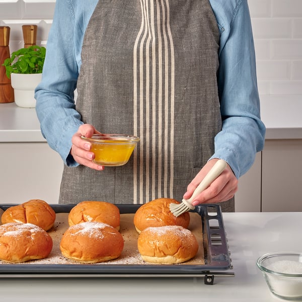 A person brushes glaze onto baked goods on a tray using a pastry brush. The tray is on a table with other baking items nearby.