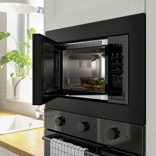 Modern kitchen scene: open black built-in microwave with food inside, above black stove with knobs, beside potted plant.