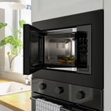 Modern kitchen scene: open black built-in microwave with food inside, above black stove with knobs, beside potted plant.