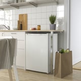 A modern white compact fridge under a kitchen counter with white cabinets and wooden floors, with a paper bag of groceries beside it.
