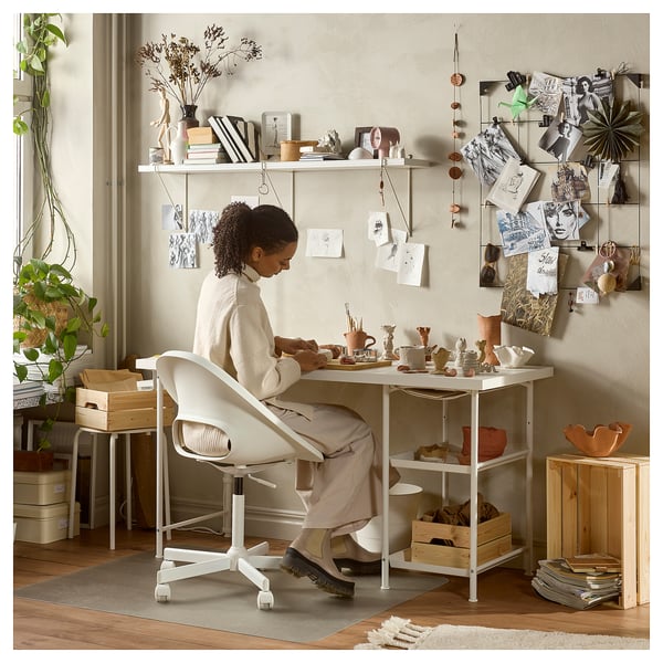 A woman works at a white LAGKAPTEN desk with shelves, surrounded by pottery and plants, with a bulletin board behind her.