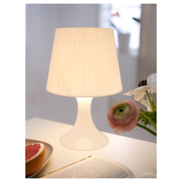 A small white lamp on a table, emitting soft light, with a pale pink flower nearby and a slice of grapefruit on a plate.