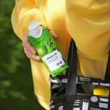 Person holding a green länglur smoothie drink carton with a white cap, wearing a yellow jacket and holding a black and white reusable shopping bag.