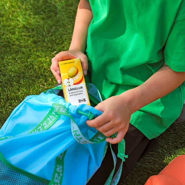 Person holding ikea LÅNGLUR fruit and vegetable smoothie box, sitting on blue mesh bag with green trim.