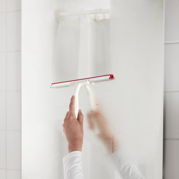 A person is cleaning a shower wall using a red-handled white squeegee, removing water and maintaining cleanliness.