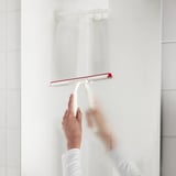A person is cleaning a shower wall using a red-handled white squeegee, removing water and maintaining cleanliness.