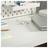 Clean, minimalist white desk setup with laptop, earphones, pencil, and shelves holding various items.