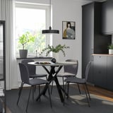 Modern dining set with round table, grey chairs, flowers, and candles. Large window and dark cabinets in background.
