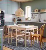 Modern kitchen with light wood dining set, green cabinets, hanging bamboo lamp, white countertops, and blue rug.
