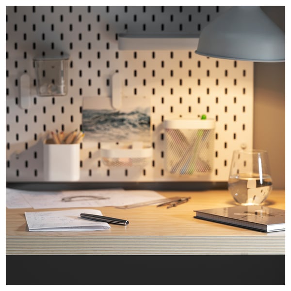 Modern desk with wooden top, blue lamp, white glass, and scattered stationery. Pegboard wall in background.