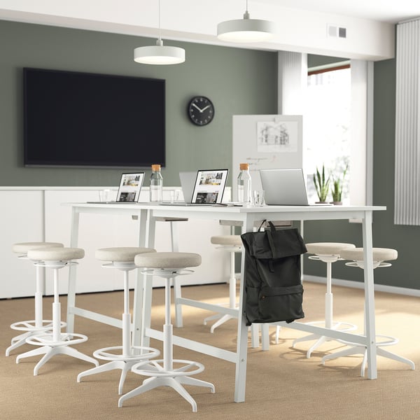 Modern office setup: high MITTZON tables with white tables & beige stools, flat screens, laptops, & backpacks.