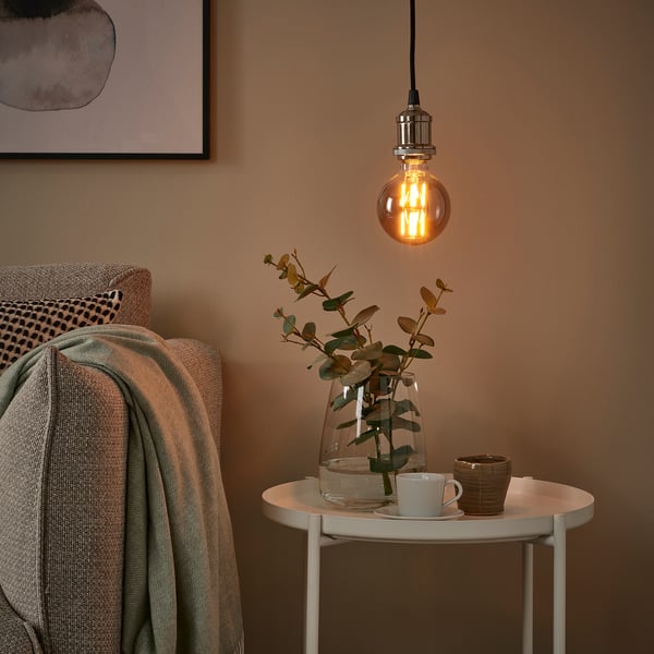Cozy corner with hanging bulb, side table, blanket, and eucalyptus vase.