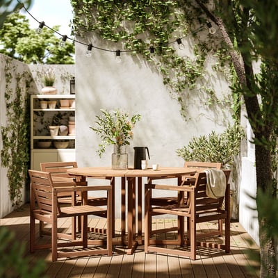 Outdoor dining set with 4 wooden chairs and table, under string lights and a white wall with greenery.