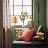 A cozy chair by a green window with an orange pillow, bowl of popcorn, lamp, and plant.