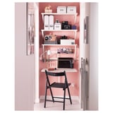 Compact home office setup with foldable black chair, pink walls, shelves with storage boxes, camera, laptop, and desk lamp.