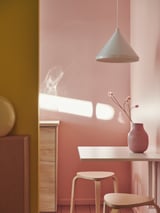 Modern dining area with pink walls, white pendant lamp, wooden table, two light wooden stools, and a red vase with flowers.
