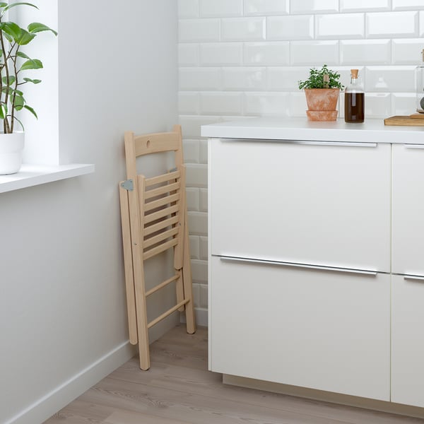 Foldable wooden chair leaned against a white wall next to a kitchen island with white cabinets and a plant on the countertop.