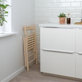 Foldable wooden chair leaned against a white wall next to a kitchen island with white cabinets and a plant on the countertop.
