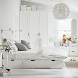 Minimalist white bedroom with NORDLI bed, storage drawers, black accents, gray pillows, plants, and large spherical light.