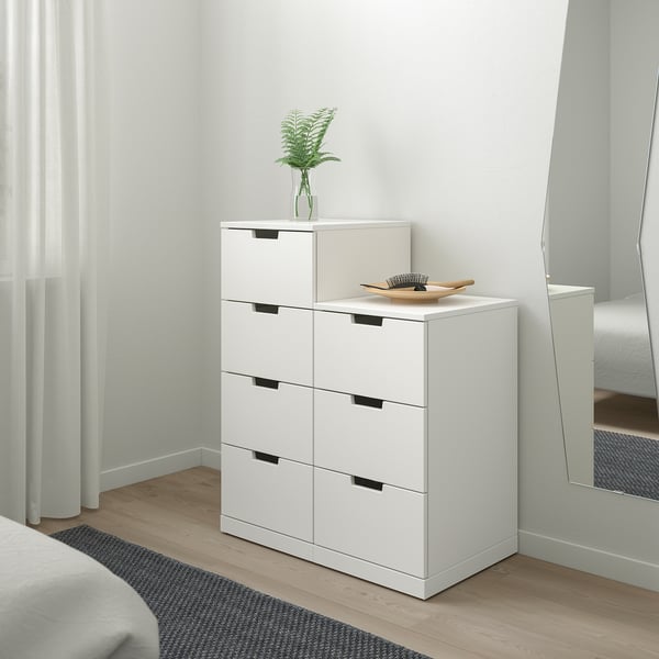 Minimalist white dresser with six drawers, plant, and bowl in bedroom, mirror reflects room.