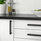 Modern kitchen with white drawers and black handles, holding a bowl of salad, plates, bottles, and a plant.